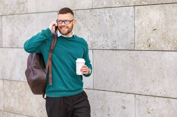 Genç, mutlu bir ofis çalışanı cep telefonuyla konuşuyor elinde kahve, şehir binasının beton duvarına karşı duruyor, dışarıda akıllı telefonlu modern girişimci.