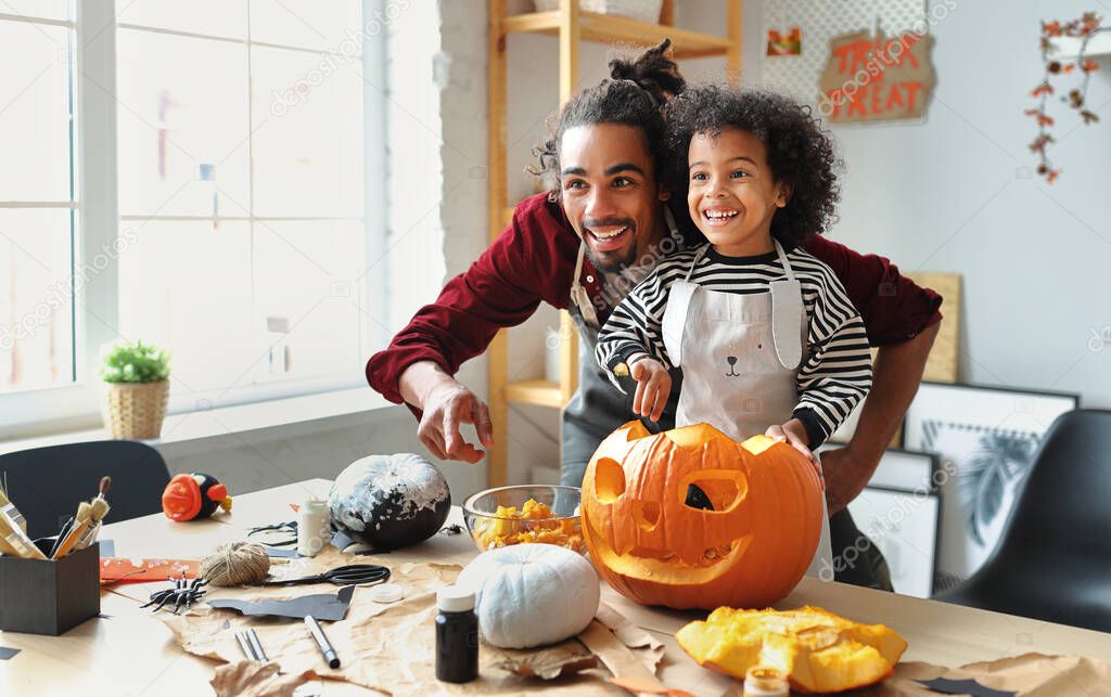 Smiling african american parent father removing pulp from ripe pumpkin while carving jack o lantern with little son for Halloween celebration at home in kitchen and looking   with smile