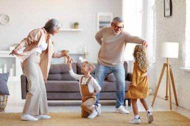 Aktif kıdemli büyükanne ve büyükbaba, oturma odasında iki mutlu torunla dans ediyor, küçük çocuklar, kız kardeş ve erkek kardeş oturma odasında büyük anne ve büyükbabayla oynarken eğleniyorlar.