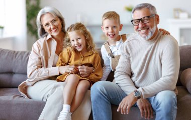 Mutlu aile, neşeli küçük çocuklar, büyük anne ve büyük babalarıyla kucaklaşırken evdeki oturma odasında birlikte oturuyorlar. Neşeli büyükanne ve büyükbaba kameraya gülümsüyorlar.