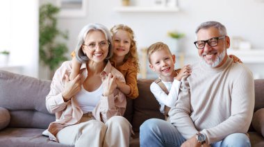 Mutlu aile, neşeli küçük çocuklar, büyük anne ve büyük babalarıyla kucaklaşırken evdeki oturma odasında birlikte oturuyorlar. Neşeli büyükanne ve büyükbaba kameraya gülümsüyorlar.