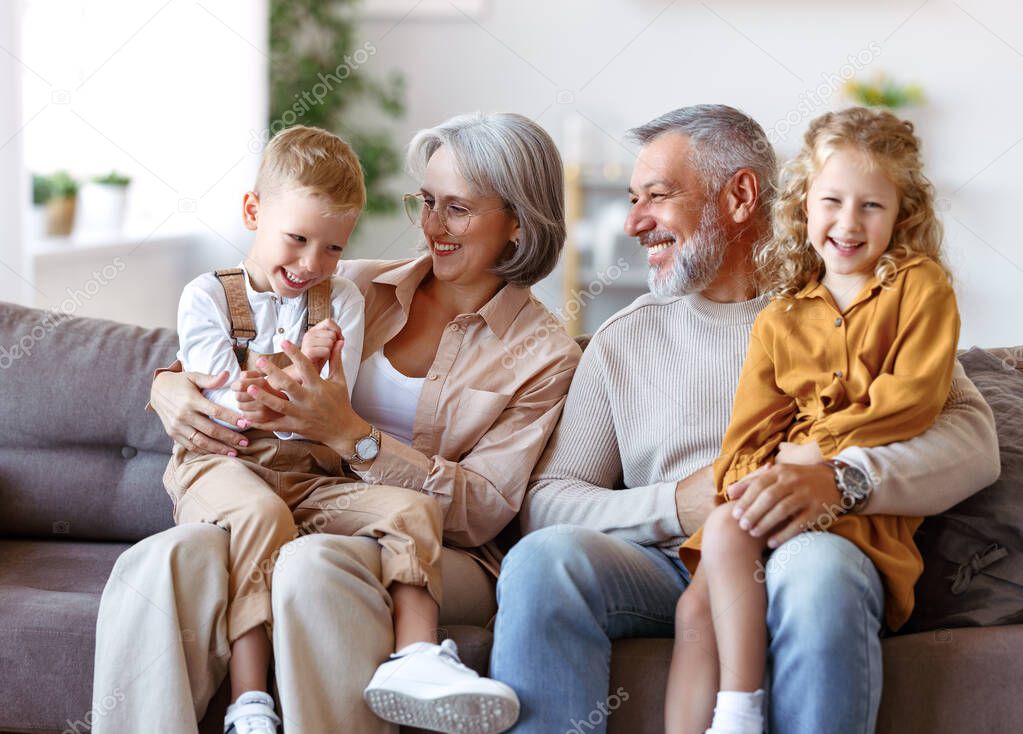 Happy family joyful little children hugging embracing with positive senior grandparents while sitting together on sofa in living room at home, cheerful grandma and grandpa with kids smiling at camera