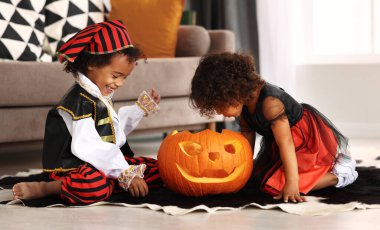 Mutlu Afrikalı küçük çocuk ve kız Cadılar Bayramı kostümleri giymiş yerde oturmuş büyük balkabağı ile oynuyorlar. Çocuklar Azizler Günü 'nü kutlarken oyulmuş balkabağının içine bakıyor.