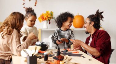 Genç, neşeli Afrikalı Amerikalı anne, baba ve iki çocuk Cadılar Bayramı için evde hazırlanıyor, balkabağı boyuyor ve Azizler Günü için kağıt kesme ve süsleme yapıyor.