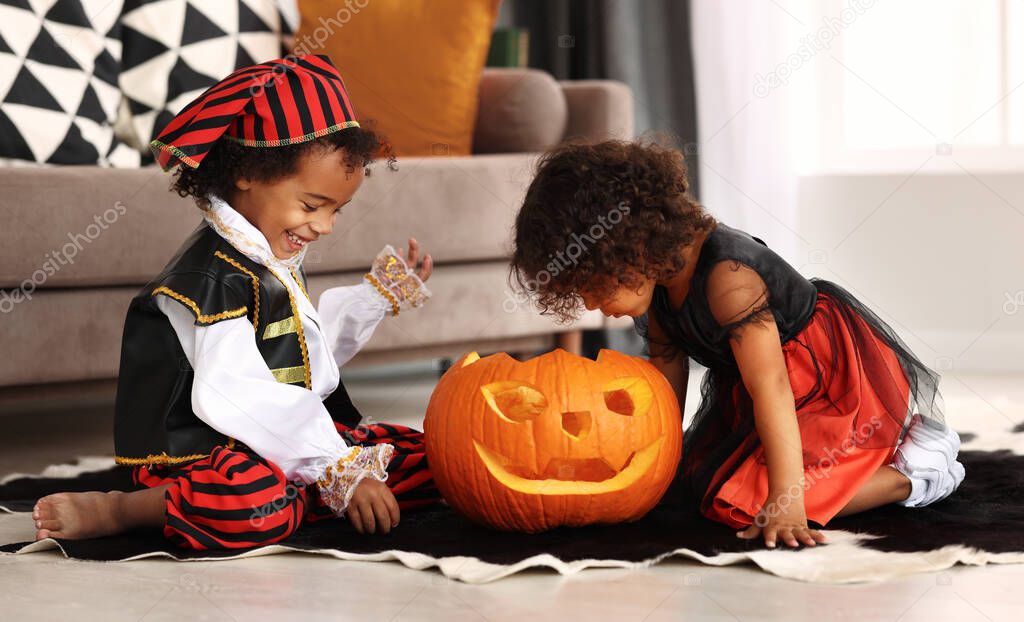 Happy excited african little boy and girl wearing Halloween costumes sitting on floor and playing with big jack-o-lantern, children looking inside of carved pumpkin while celebrating All Saints Day