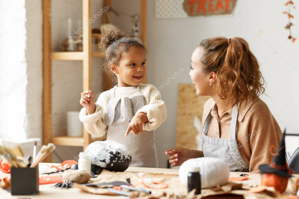 Cute little african american girl holding paintbrush and painting Halloween pumpkin with mom together at home, loving parent and kid sitting at table and preparing handmade decorations for Saints Day party