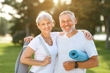 Smiling senior couple with exercise mats standing at park, positive mature man embracing his elderly wife after fitness or yoga class in nature. Wellness and healthy lifestyle on retirement clipart