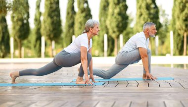 Mutlu, olgun, olgun bir çift yaşlı kadın ve erkek yogası yapıyor dışarıda açık havada çıplak ayakla, Virabhadrasana pozu veriyor. Şehir parkının arka planında, çıplak ayakla.