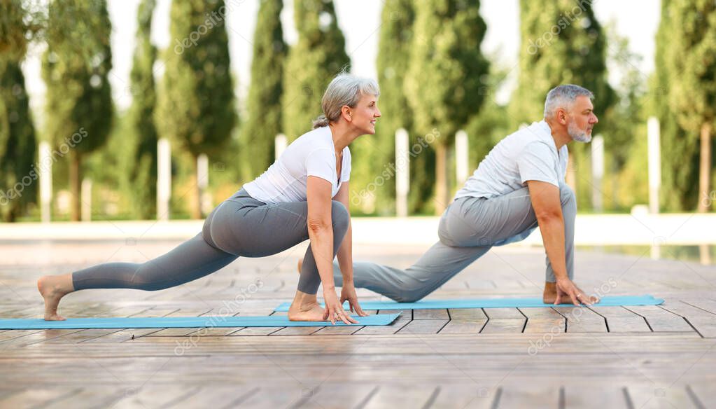 Happy positive fit mature couple of senior woman and man practicing partner yoga on open fresh air outside standing barefoot in warrior Virabhadrasana pose against background of city park exterior
