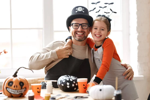 Portrait of happy smiling family dad and daughter wearing Halloween hats smiling at camera and embracing while preparing handmade home decorations, father and little girl enjoying painting pumpkins