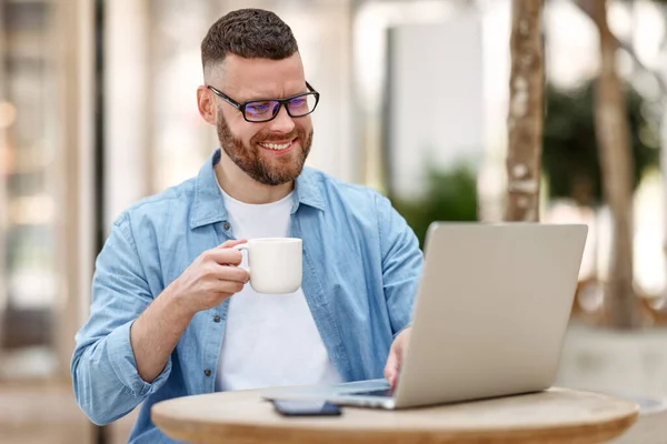 Joven hombre feliz freelancer trabajando remotamente en el ordenador portátil, beber café mientras está sentado en la cafetería al aire libre — Foto de Stock