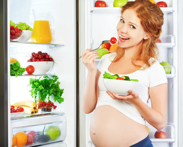 Feliz mujer embarazada comiendo ensalada cerca del refrigerador —  Fotos de Stock