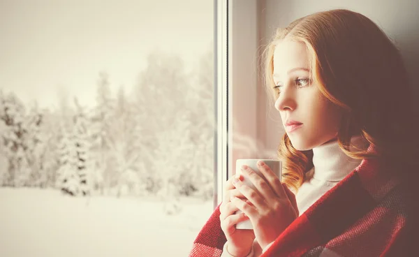 Pensive fille triste avec une boisson chauffante regardant par la fenêtre dans — Photo