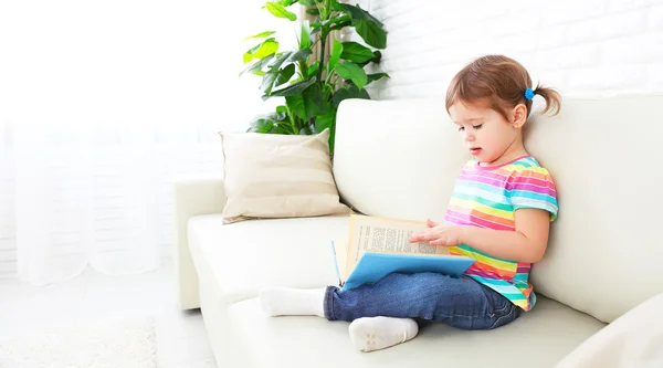 Мила дівчинка читає книгу вдома, сидячи на дивані — стокове фото