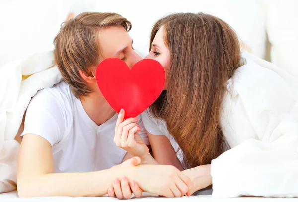 Amante pareja besándose con un corazón rojo en la cama —  Fotos de Stock