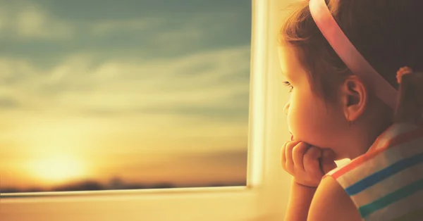 Enfant triste petite fille regardant par la fenêtre au coucher du soleil — Photo