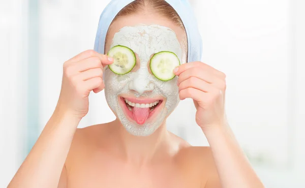 Chica joven divertida con una máscara para la cara de la piel y los pepinos — Foto de Stock