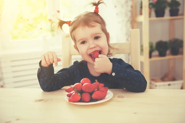 Happy child girl eats strawberries in summer home kitchen — Stock Photo, Image