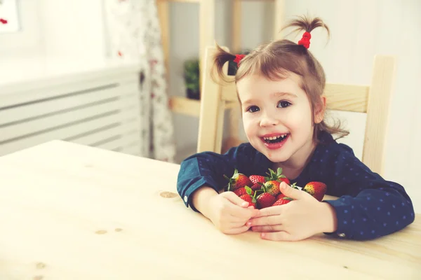 Happy child girl eats strawberries in summer home kitchen — Stock Photo, Image