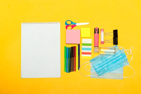 Back to school, school supplies with medical face mask and empty notebook on a yellow background. Protection of schoolchildren and students from the virus
