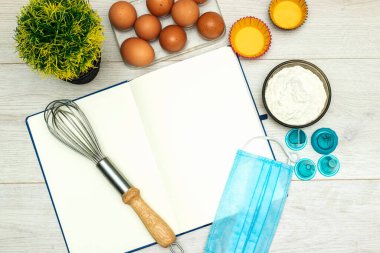 Boş tarif kitabını aç, yumurta ve un, ahşap masada tıbbi maske.