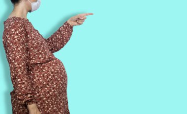 Tıbbi maske takan ve parmağını bir şeye doğrultan hamile bir kadın.