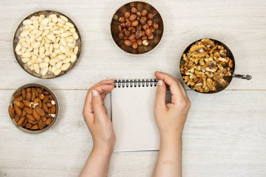 Metal kaselerde fındıklar, cevizler, bademler ve boş bir defter tutan eller.