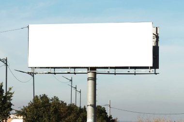 Büyük boş bir reklam panosu, açık hava ortamında uzun bir direğin tepesinde duruyor. Parlak mavi bir gökyüzü, elektrik hatları ve arka planda reklam ya da sanat için hazır ağaçlar var.