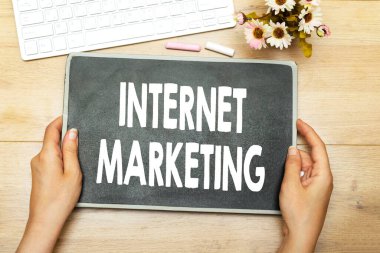 İnternet pazarlaması yazan karatahta iki elle ahşap bir masada tutulur. Yakınlarda bir klavye ve küçük bir buket dijital pazarlama fikirleri için parlak ve verimli bir çalışma alanı oluşturur.