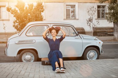 Gülümseyen kadın şık, eski moda bir arabanın yanında poz veriyor. Bavulun üzerinde oturan kadın. Sokak tarzı ve şehir yürüyüşü konsepti