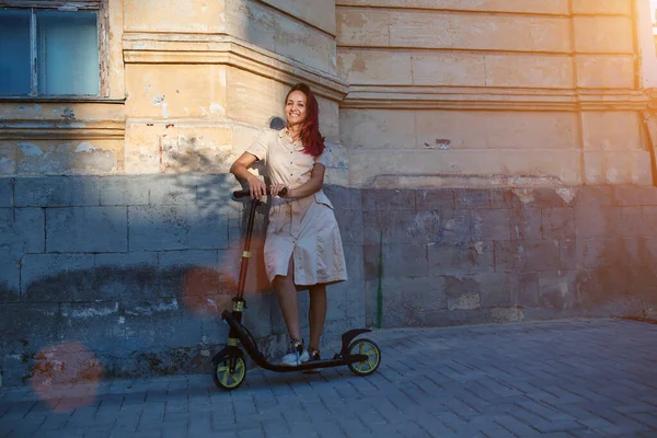 Счастливая красотка в платье со скутером. Солнце светит на фасад здания. Активный отдых и новая идея места, копировальное пространство — стоковое фото