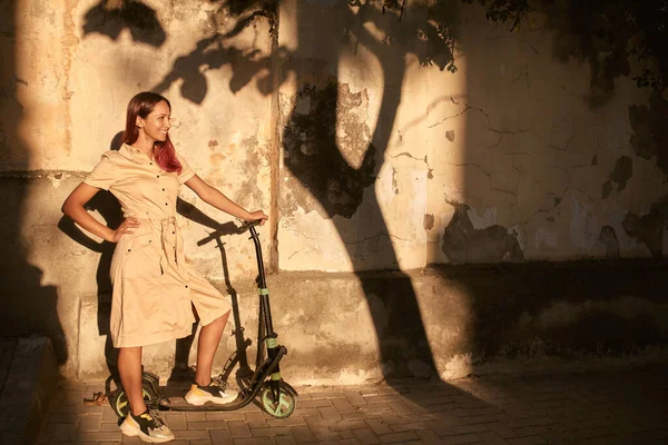 Улыбающаяся девушка позирует возле стены со скутером. Солнечный свет падает на старую стену. Новые эмоции и идея активного отдыха. — стоковое фото
