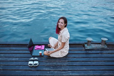 Serbest meslek ya da uzaktan öğrenme konsepti. Bayan dizüstü bilgisayarı ve not defteriyle suya yakın oturuyor. Seyahat ve öğrenim fikri