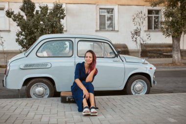 Güzel kız bavulun üzerinde oturuyor ve gülümsüyor. Mavi klasik arabanın yanında poz veren bir kadın. Yeni yerler ve yeni duygu kavramı