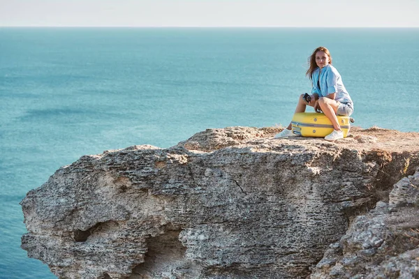 Leuke vrouw die poseert op een gele koffer. Prachtig uitzicht op de oceaan vanaf de berg. Nieuw terrein en het bereiken van doel idee, kopieer ruimte — Stockfoto