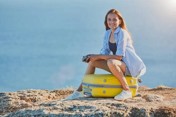 Smiling pretty lady posing on yellow luggage. Ocean view on background. Business travel and new ground idea, copy space — Stock Photo, Image