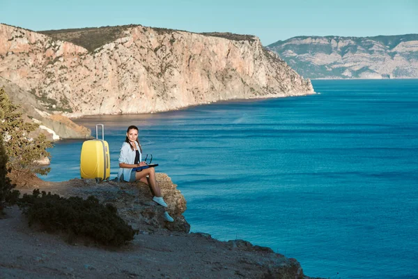 Remote office and business travel concept, copy space. Cute lady work sitting on rock. Bright yellow luggage for trip — Stock Photo, Image