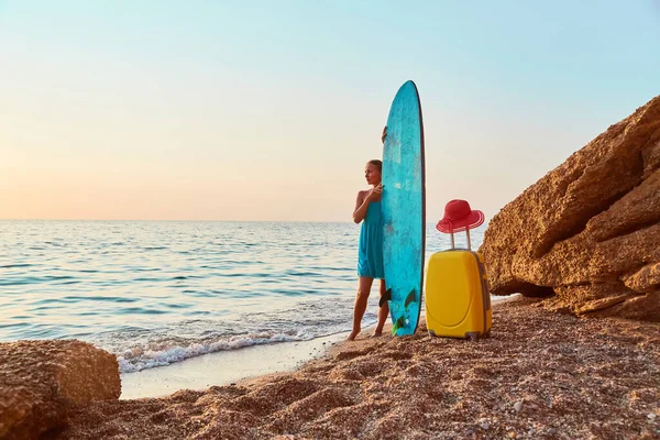 Жінка насолоджується видом на океан, тримаючи дошку для серфінгу, багаж і капелюх на піску. Літній час, відпустка та спортивна ідея серфінгу — стокове фото