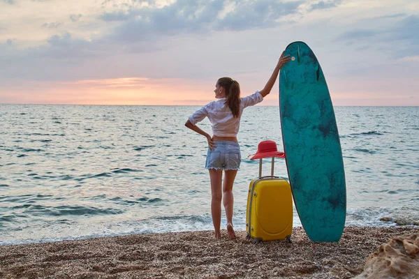 Femme en vacances, planche de surf et valise. Coucher de soleil sur l'océan, sens de l'aventure et concept de temps de détente. Espace de copie Photos De Stock Libres De Droits