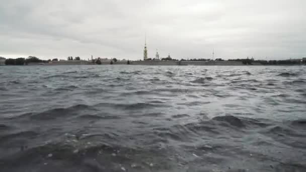 Petropavlovskaja fort en de Neva rivier. Actie. St. Petersburg, Rusland, close-up van rimpelwater oppervlak van een rivier en de vesting ver in de verte op bewolkte lucht achtergrond. — Stockvideo