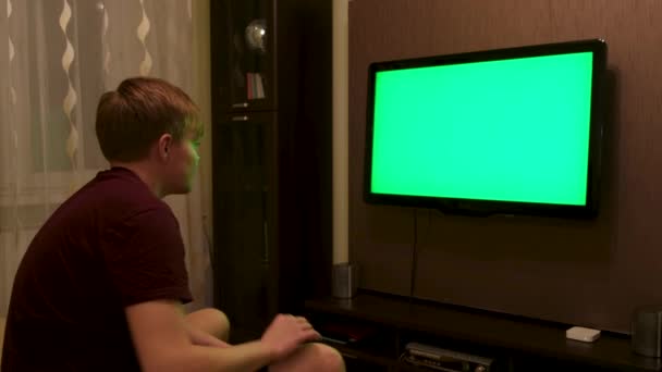 Muž sedí na gauči v obývacím pokoji a dívá se na televizi se zelenou obrazovkou. Koncept. Boční pohled na mladého muže, jak vypíná televizi s klíčem chroma a odchází. — Stock video