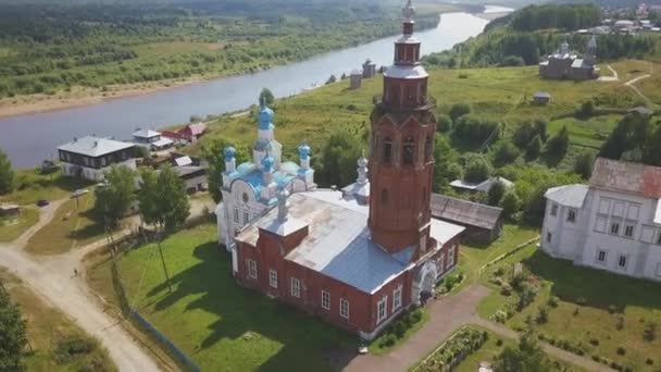 Vista aérea de una antigua iglesia en el pequeño pueblo. Clip. Paisaje rural de verano con un río y un pueblo cerca de iglesia blanca y campanario de ladrillo rojo. — Vídeo de stock