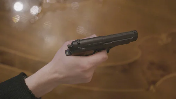 Close-up of hand holding gun at gunpoint. Stock. Dangerous criminal threatens with gun on background of wooden table. Criminal with gun threatens rich people