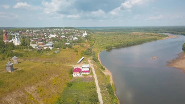 Top view of green town by river in summer. Clip. Picturesque panorama with cottage town and river in cloudy weather. Beautiful town located on plain by river