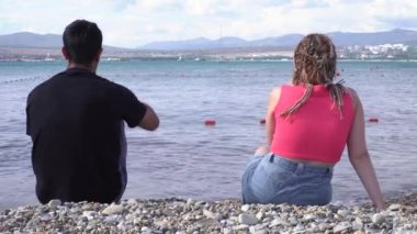 Kadın ve erkek, deniz kıyısındaki çakıl taşı plajında otururken ellerinde kırmızı şarap kadehleri tutuyorlar. Medya. Sahilde romantik bir çiftin arka görüntüsü..