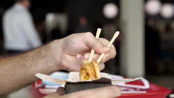 Close-up van mannelijke handen met houten Chinese eetstokjes die heerlijke Aziatische wok noedels eten. De media. Zijaanzicht van het eten van Chinese pasta op een openbare plaats. — Stockvideo