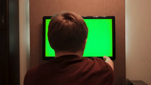 Rear view of man looking at green TV. Concept. Man watches TV with green screen and presses remote control. Man switches channels on TV with green screen