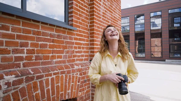 Женщина-фотограф смеется над уличной фотосессией. Начали. Красивая женщина держит профессиональную камеру и смеется на фоне кирпичной стены — стоковое фото