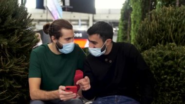 Erkek arkadaşlar akıllı telefon cihazıyla eğleniyor. Medya. İki erkek arkadaş yeşil çalıların yanında oturmuş sohbet ederken, gülerken ve akıllı telefonlara bakarken tıbbi maske takıyorlar..