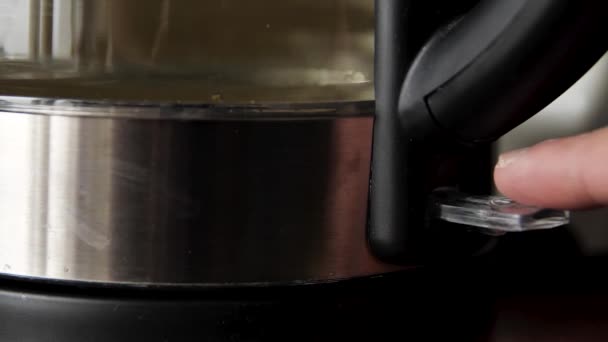 Мужской палец нажимает выключатель на электрическом чайнике. СМИ. Закрытие процесса нагрева чистой питьевой воды в чайнике с синим освещением. — стоковое видео